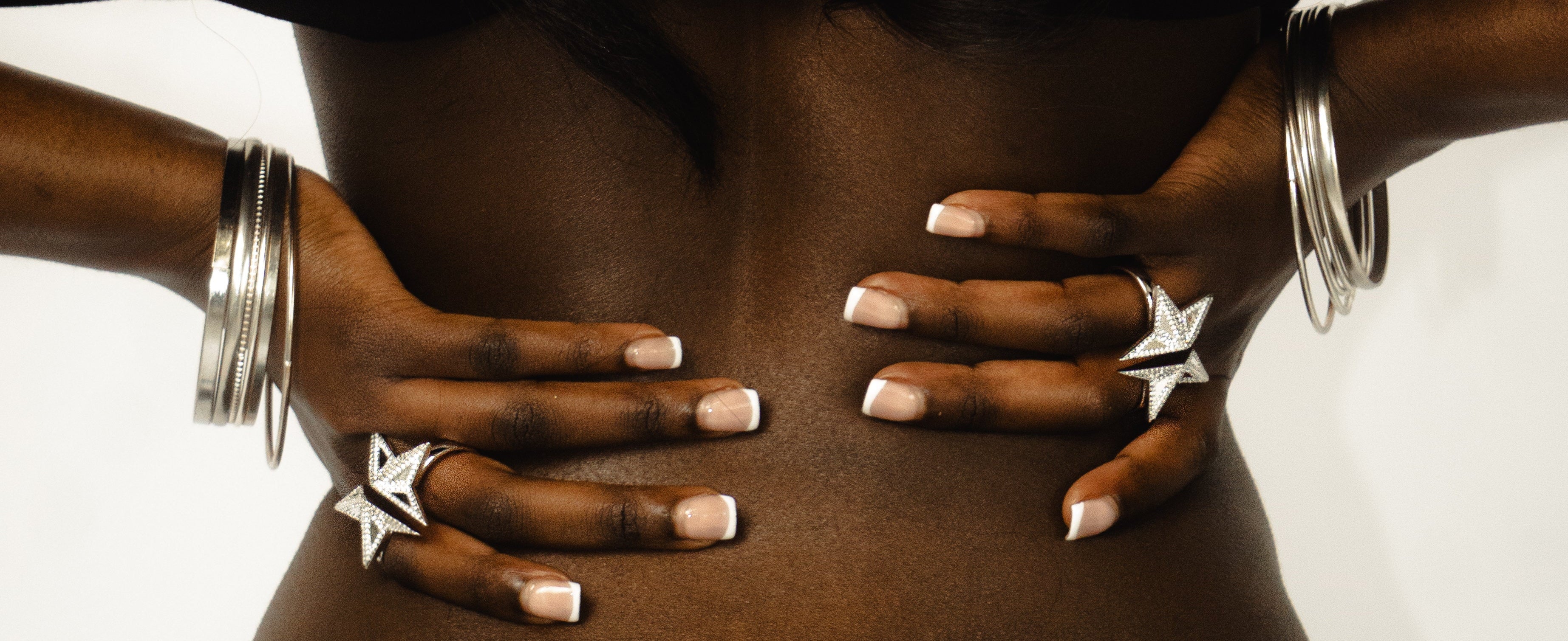 A close-up lifestyle shot of a person's bare back and hands, wearing multiple silver star-shaped rings and stacked silver bangles against a plain white background.